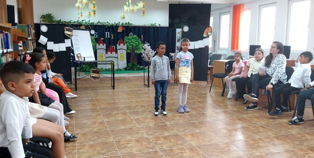 Момче и момиче стоят в центъра на залата пред седнали деца по време на занимание в библиотека.