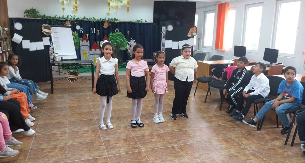 Четири момичета стоят в редица пред други деца по време на събитие в библиотека.